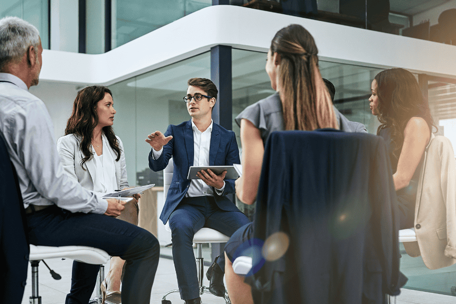 a man, holding a tablet, speaks to a group of five professionals sitting in a circle