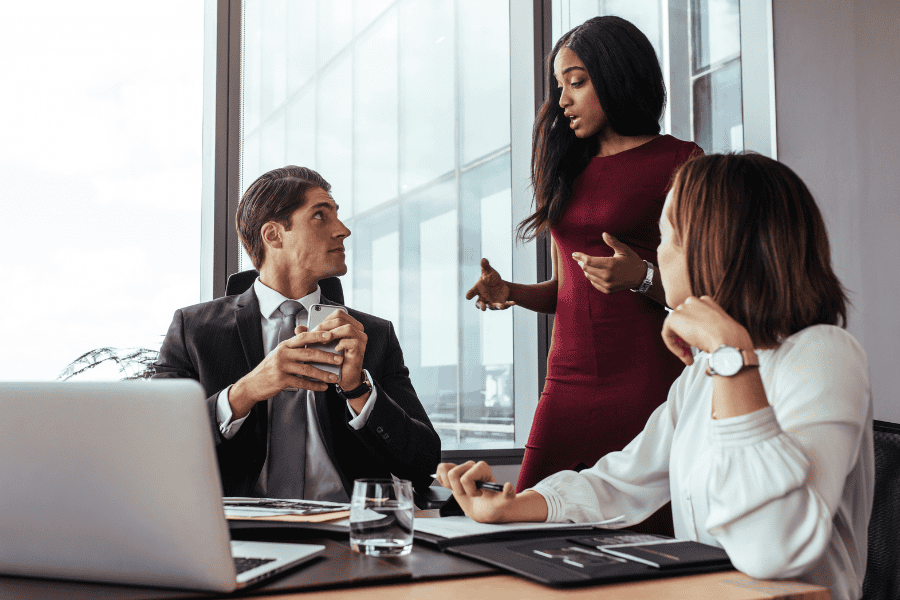 A team of three business professionals engaged in a discussion around a desk in a modern office.