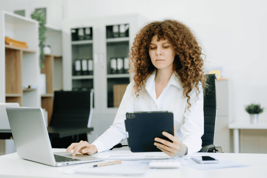 a professional woman checking HMRC ERS filings on a laptop while holding a tablet in an office