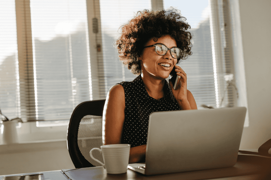 Smiling woman with curly hair using phone and laptop in office.